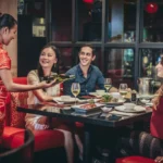 Waitress in red cheongsam serving wine to smiling diners at an elegant Asian restaurant. Group enjoying dinner with food and drinks.