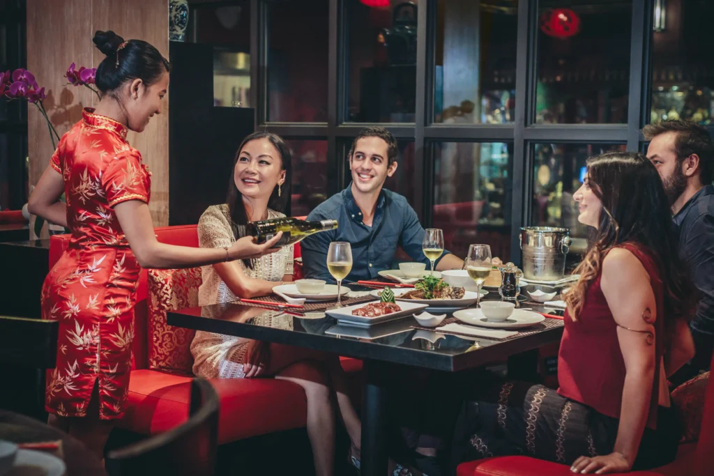 Waitress in red cheongsam serving wine to smiling diners at an elegant Asian restaurant. Group enjoying dinner with food and drinks.