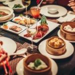 A vibrant table spread of various Chinese dishes including dim sum, roasted duck, and entrees, with diners enjoying the meal.