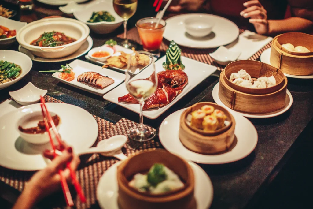 A vibrant table spread of various Chinese dishes including dim sum, roasted duck, and entrees, with diners enjoying the meal.