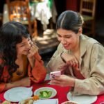 Two smiling women at a restaurant table looking at a smartphone, sharing a moment. Food and drinks are on the red tablecloth.