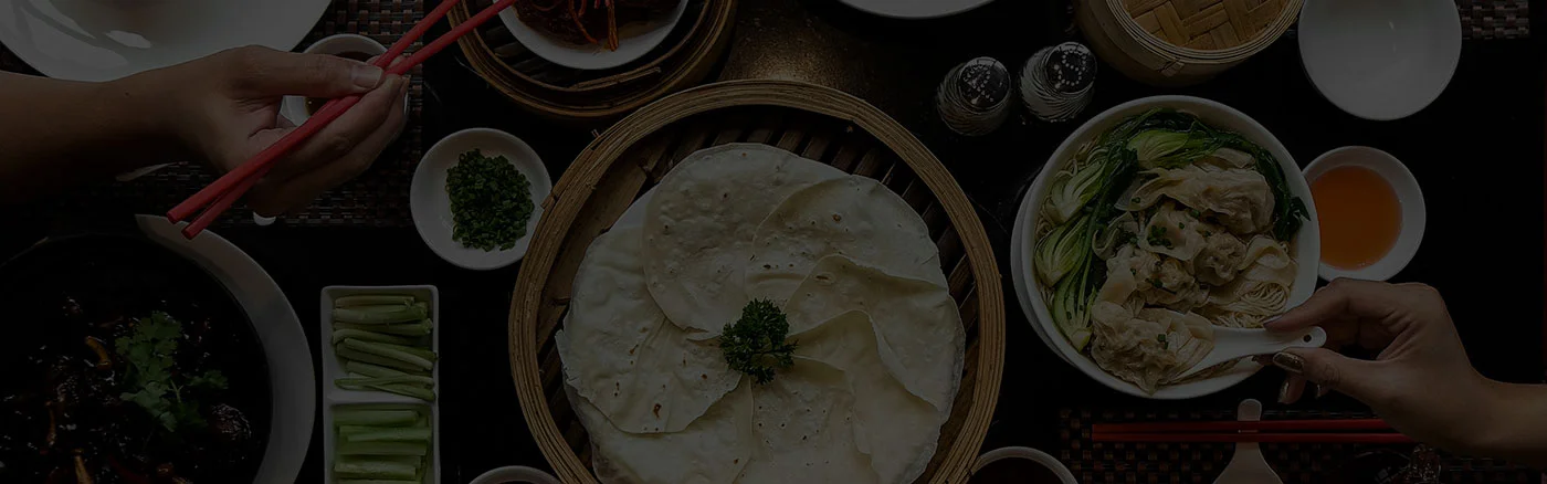 Overhead view of a table laden with various Asian dishes, including wonton noodle soup, pancakes in a steamer, and sides, with hands using chopsticks and a spoon.