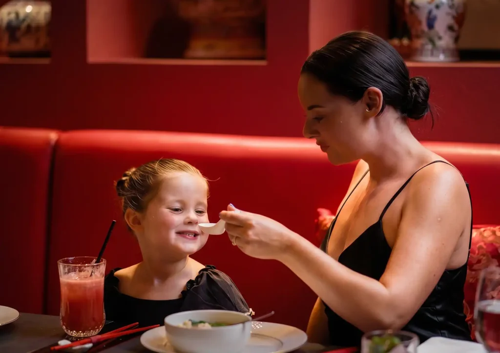 Best dishes for children at family restaurant — smiling child fed soup with spoon, pink drink and chopsticks on table.