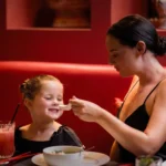 Best dishes for children at family restaurant — smiling child fed soup with spoon, pink drink and chopsticks on table.