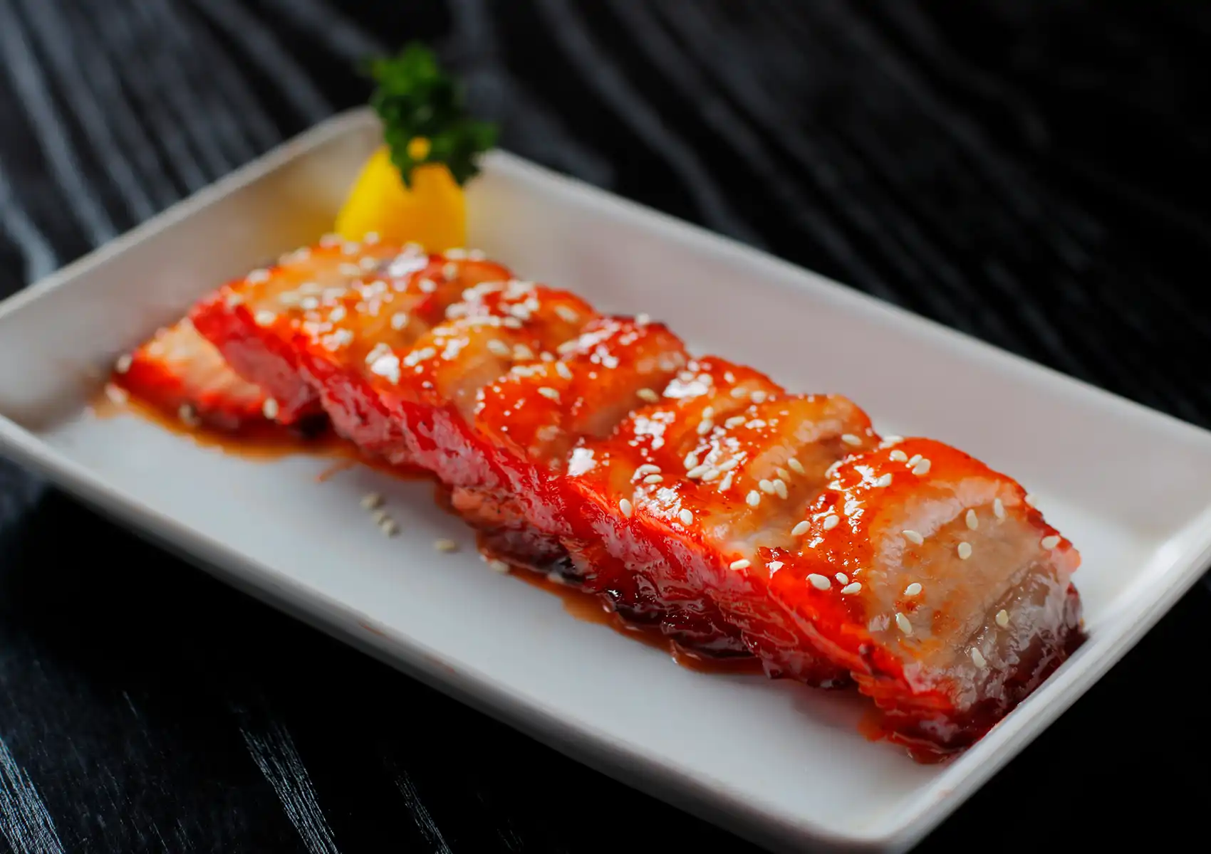 Chinese char siu barbecue pork slices with sesame seeds, lemon wedge, and parsley garnish on white plate.