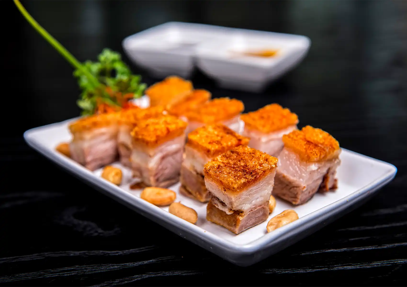 Crispy roasted pork belly slices with golden skin, peanuts, dipping sauces, and fresh herb garnish on white plate.