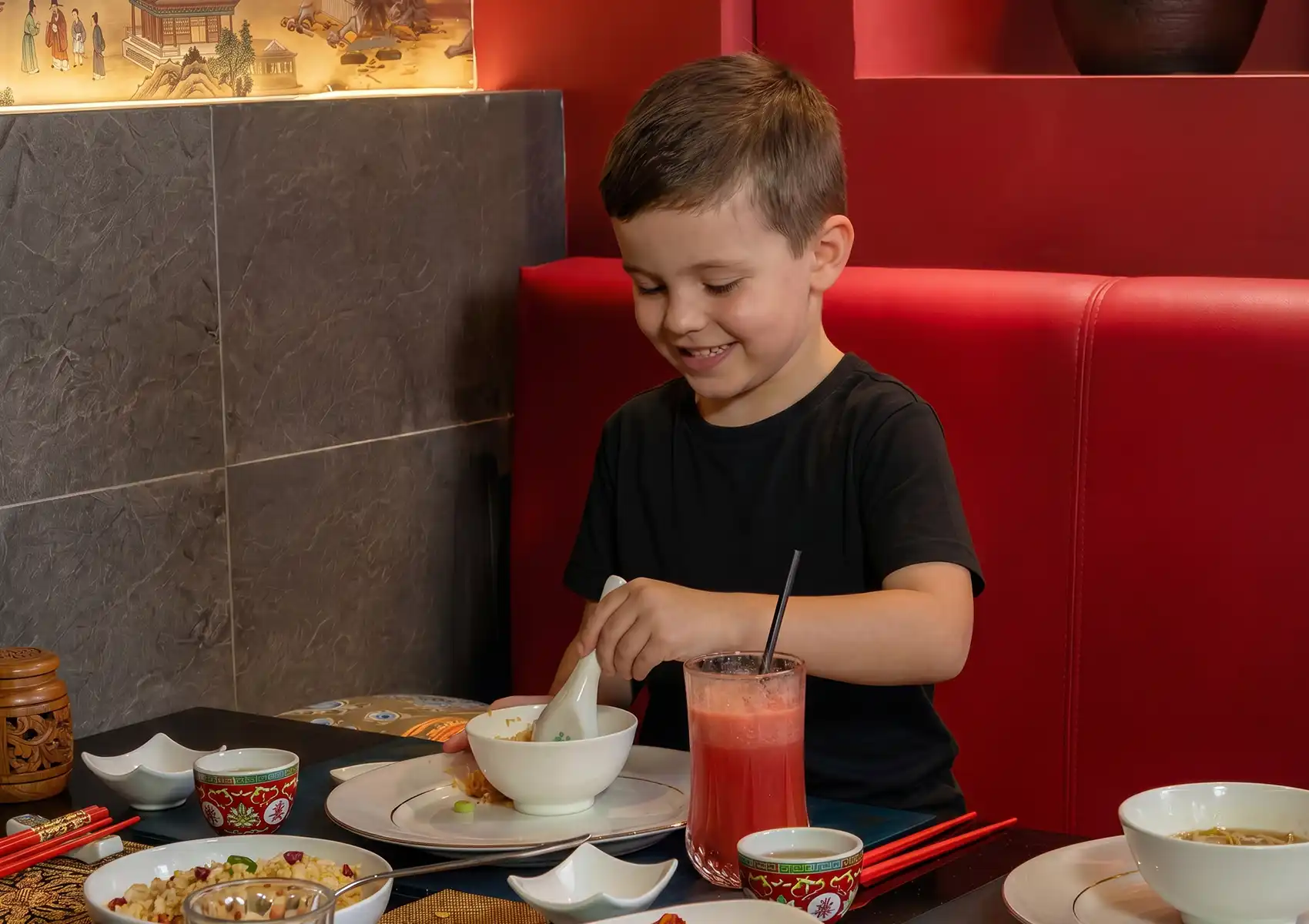 Smiling child enjoying meal at Asian restaurant table with rice, soup, pink drink, and chopsticks.