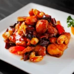 A white square plate of kung pao chicken at a Cantonese restaurant in Bali, with glazed chicken pieces, cashew nuts, dried red chilies, and bell peppers in a rich sauce, garnished with parsley.