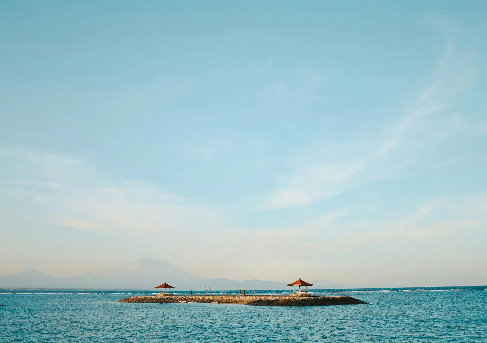 A calm ocean view in Sanur with traditional Balinese gazebos on a stone jetty and distant mountains, near a Cantonese restaurant in Bali.