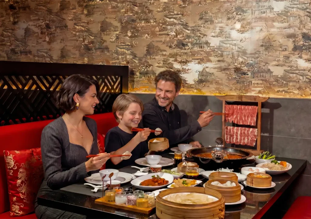 A family enjoying hot pot and dim sum together in a booth at a Cantonese restaurant in Bali, with sliced meats, bamboo steamers, and shared dishes spread across the table.