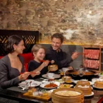 A family enjoying hot pot and dim sum together in a booth at a Cantonese restaurant in Bali, with sliced meats, bamboo steamers, and shared dishes spread across the table.