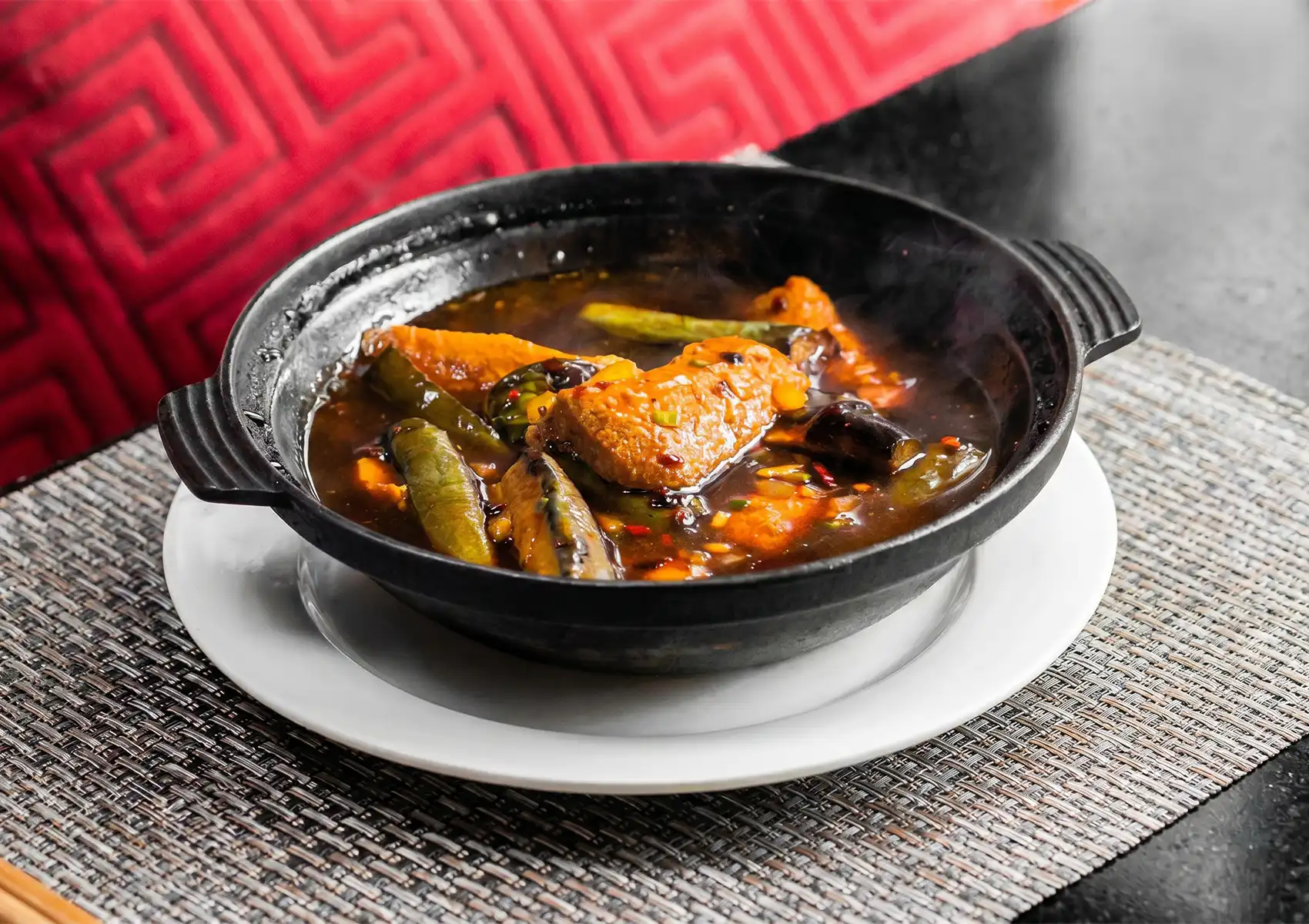 A sizzling eggplant claypot with tofu, green chilies, and aromatic brown sauce served on a woven mat at a Cantonese restaurant in Bali.