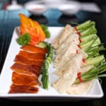 A white plate of Peking duck at a Cantonese restaurant in Bali, with crispy sliced duck skin, thin pancakes, cucumber, spring onions, and a carved tomato garnish.
