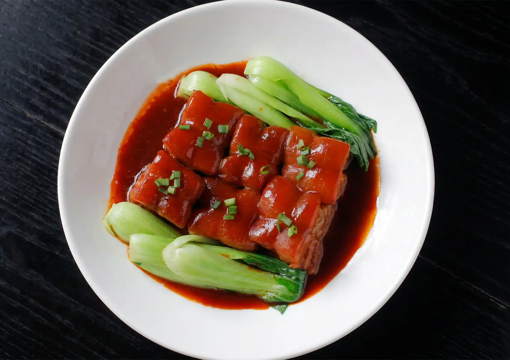 An overhead view of a beautifully plated dish featuring neatly arranged squares of glazed braised pork belly and steamed bok choy in a white bowl, showcasing the authentic culinary offerings of a Cantonese restaurant in Bali.