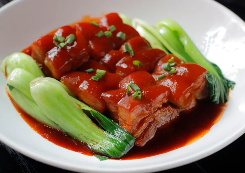 A close-up of tender braised pork belly coated in a rich, dark sauce, garnished with scallions and served alongside fresh green bok choy, highlighting a signature dish at a Cantonese restaurant in Bali.