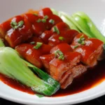 A close-up of tender braised pork belly coated in a rich, dark sauce, garnished with scallions and served alongside fresh green bok choy, highlighting a signature dish at a Cantonese restaurant in Bali.