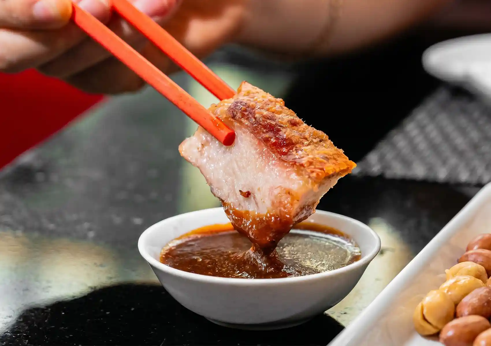 A close-up of orange chopsticks dipping a thick, succulent slice of crispy pork belly into a small bowl of rich brown sauce at a Cantonese restaurant in Bali.