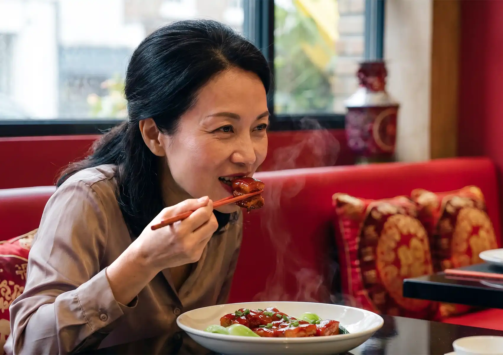 A smiling woman uses chopsticks to enjoy a steaming piece of glazed pork belly while dining in a vibrant red booth at a Cantonese restaurant in Bali.