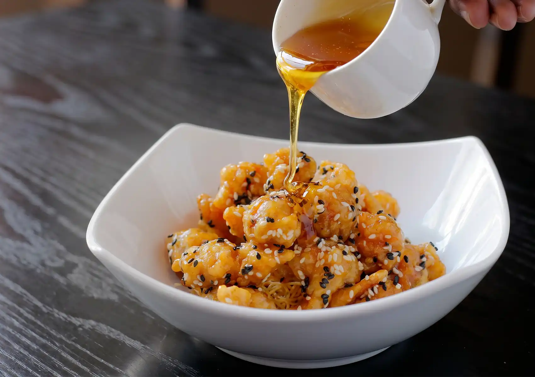 A hand carefully pours a rich, golden honey glaze from a small white pitcher over a delicious bowl of crispy sesame chicken, showcasing a mouthwatering dish available at a Cantonese restaurant in Bali.