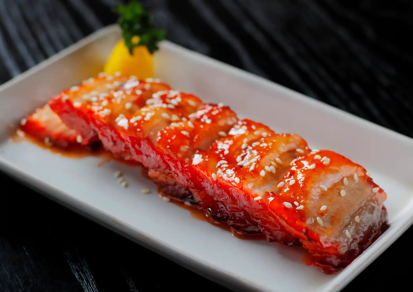 Close-up of tender, sliced char siew pork with a glossy red glaze and a sprinkle of white sesame seeds on a rectangular white plate, showcasing authentic Cantonese BBQ Ubud.