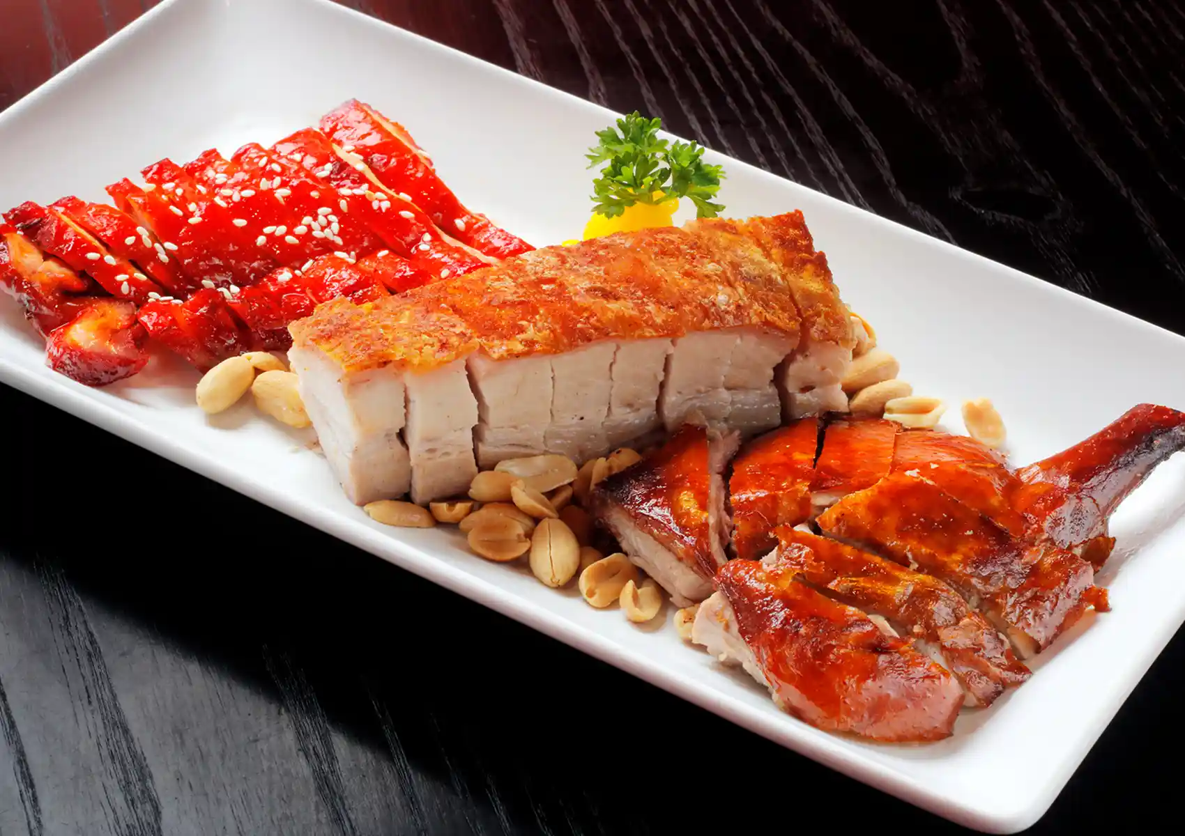 A mixed barbecue meat platter featuring bright red char siu, crispy pork belly, and glazed roast duck resting on scattered peanuts, served at a Cantonese restaurant in Bali.