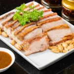 A plate of sliced crispy roasted pork belly garnished with fresh parsley, served alongside roasted peanuts and a savory dipping sauce at a Cantonese restaurant in Bali.