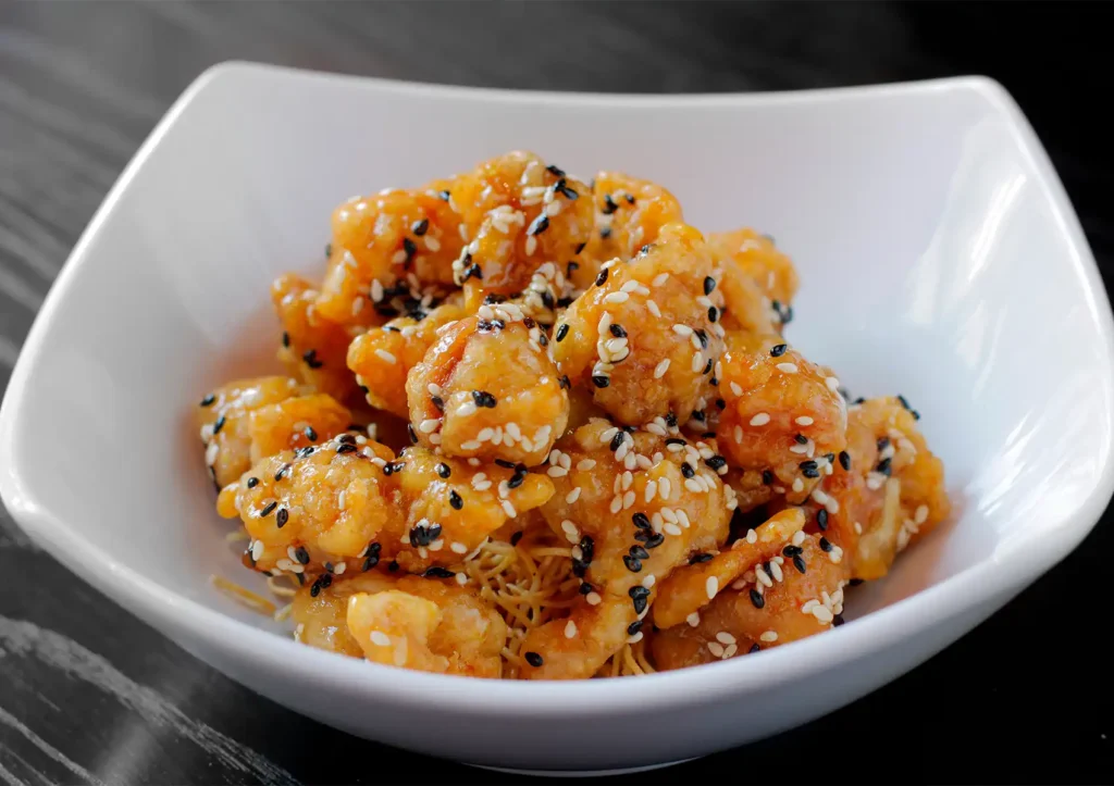 A close-up view of crispy, golden chicken pieces coated in a sweet glaze and generously sprinkled with black and white sesame seeds, served over a bed of crispy thin noodles in a white bowl at a Cantonese restaurant in Bali.