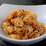A close-up view of crispy, golden chicken pieces coated in a sweet glaze and generously sprinkled with black and white sesame seeds, served over a bed of crispy thin noodles in a white bowl at a Cantonese restaurant in Bali.