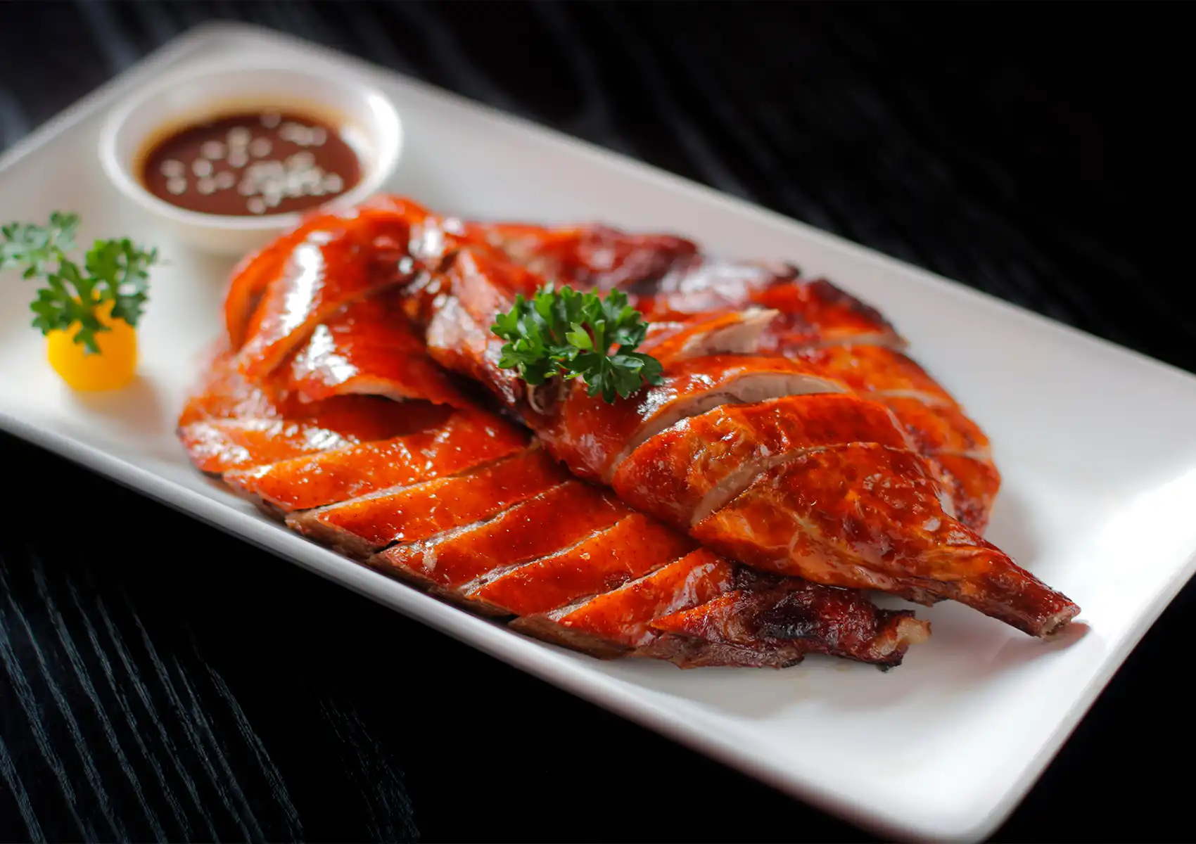 A perfectly roasted and sliced duck with crispy, golden-brown skin served on a white plate with a small bowl of dipping sauce and a parsley garnish, highlighting a delicious Cantonese BBQ Ubud dish.