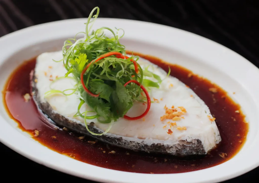 A beautifully plated fillet of steamed white fish resting in a rich soy-based broth, elegantly garnished with shredded green scallions, fresh cilantro, and red chili slivers, representing a classic seafood dish at a Cantonese restaurant in Bali.