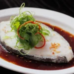 A beautifully plated fillet of steamed white fish resting in a rich soy-based broth, elegantly garnished with shredded green scallions, fresh cilantro, and red chili slivers, representing a classic seafood dish at a Cantonese restaurant in Bali.