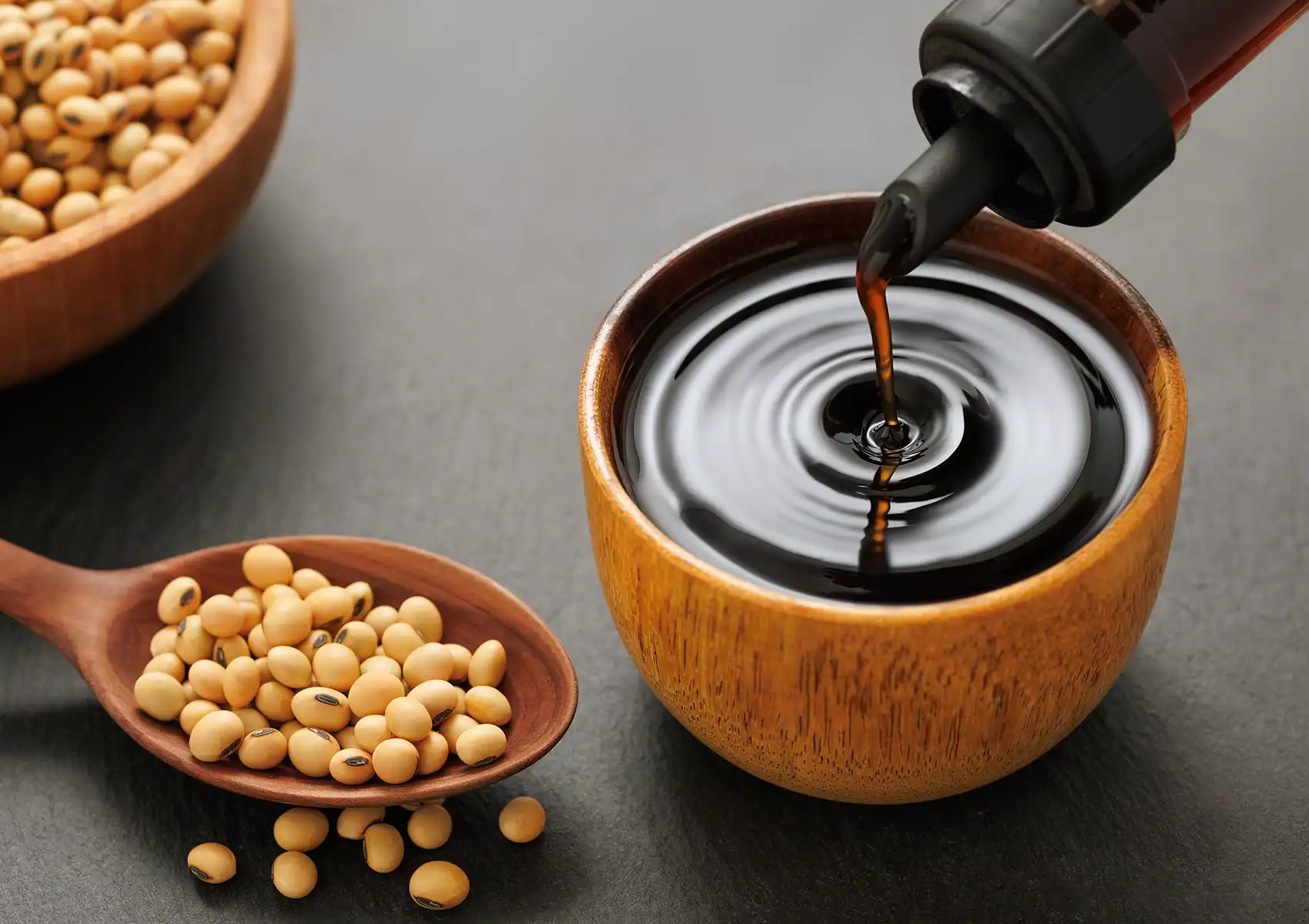 A close-up view of dark soy sauce being poured from a bottle into a small wooden bowl next to a wooden spoon filled with dry soybeans, highlighting the essential, authentic ingredients used at a Cantonese restaurant in Bali.