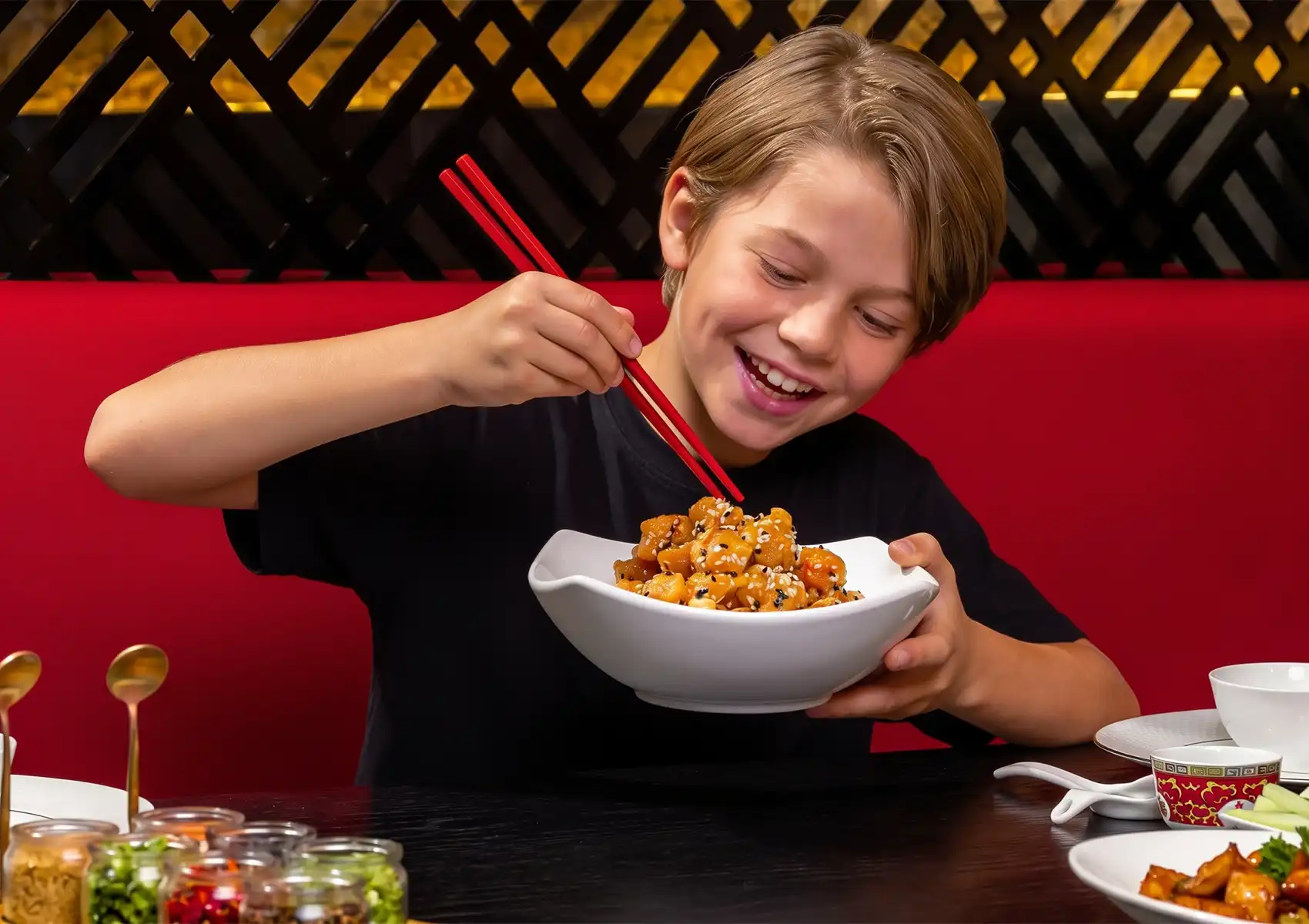 A happy young boy smiles brightly as he uses red chopsticks to eat a bowl of sweet and savory honey sesame chicken while sitting in a vibrant red booth at a Cantonese restaurant in Bali.