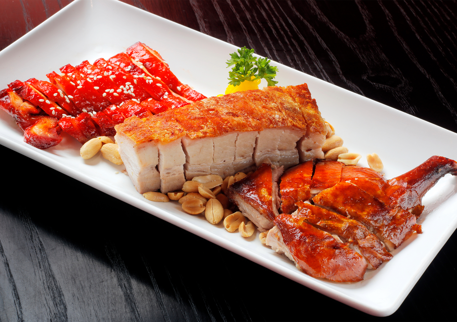 A vibrant combination platter of traditional roasted meats, featuring sweet red glazed char siu, crispy pork belly, and shiny roast duck, arranged on a white rectangular plate at a Cantonese restaurant in Bali.