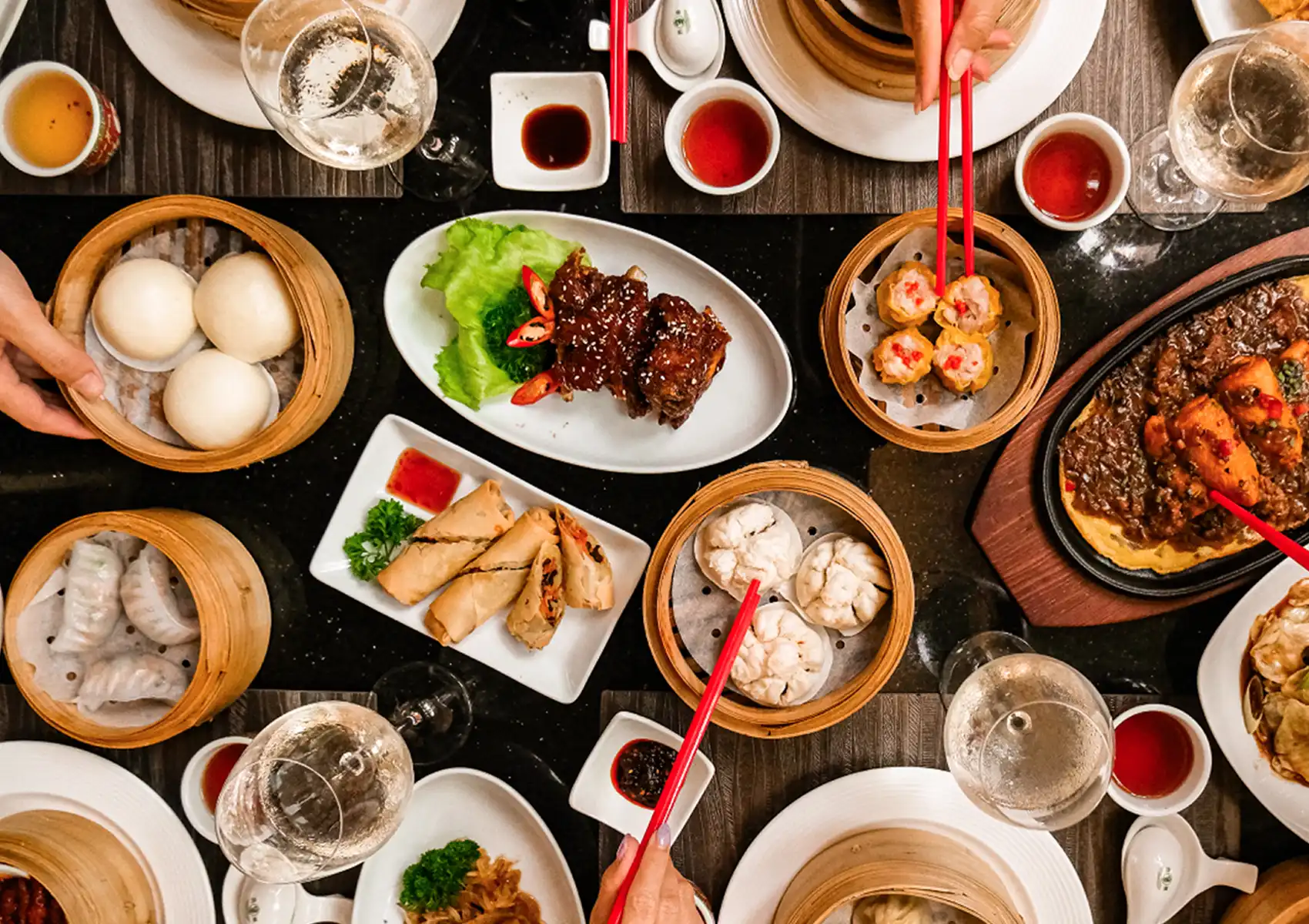 An overhead view of an abundant feast featuring bamboo steamers filled with dumplings, crispy spring rolls, and glazed ribs being shared by diners using red chopsticks at a Restaurant in ubud.