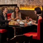 Three women happily conversing and sharing a dim sum meal with chopsticks while seated in a cozy red booth at a Restaurant in ubud.