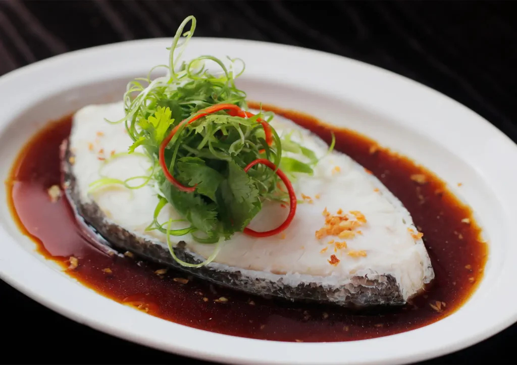 A close-up of a perfectly steamed white fish fillet resting in a rich soy-based broth, elegantly garnished with curled scallions, fresh cilantro, and red chili on a classic white oval plate at a Cantonese restaurant in Bali.