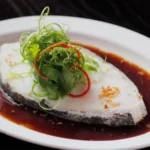 A close-up of a perfectly steamed white fish fillet resting in a rich soy-based broth, elegantly garnished with curled scallions, fresh cilantro, and red chili on a classic white oval plate at a Cantonese restaurant in Bali.