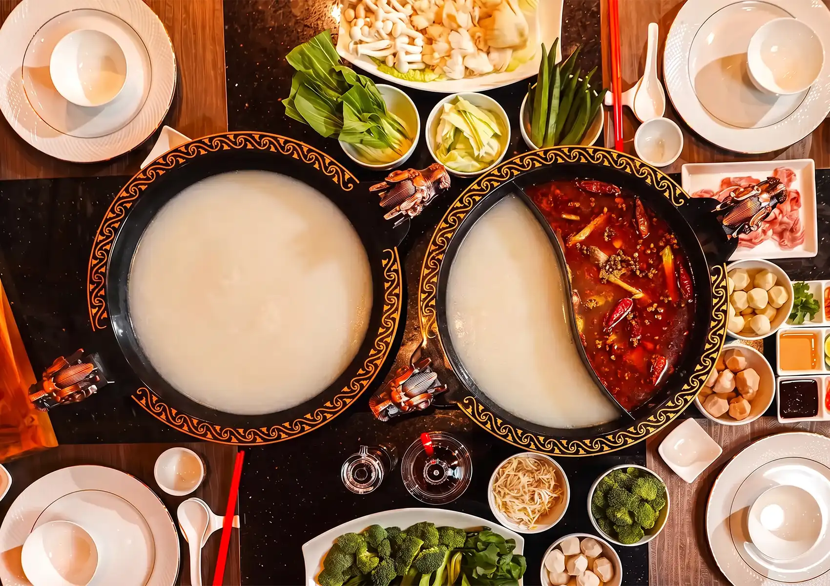 An overhead view of a lavish hot pot table spread featuring dragon-handled pots filled with creamy collagen and spicy red mala broths, surrounded by fresh vegetables and ingredients at a Cantonese restaurant in Bali.