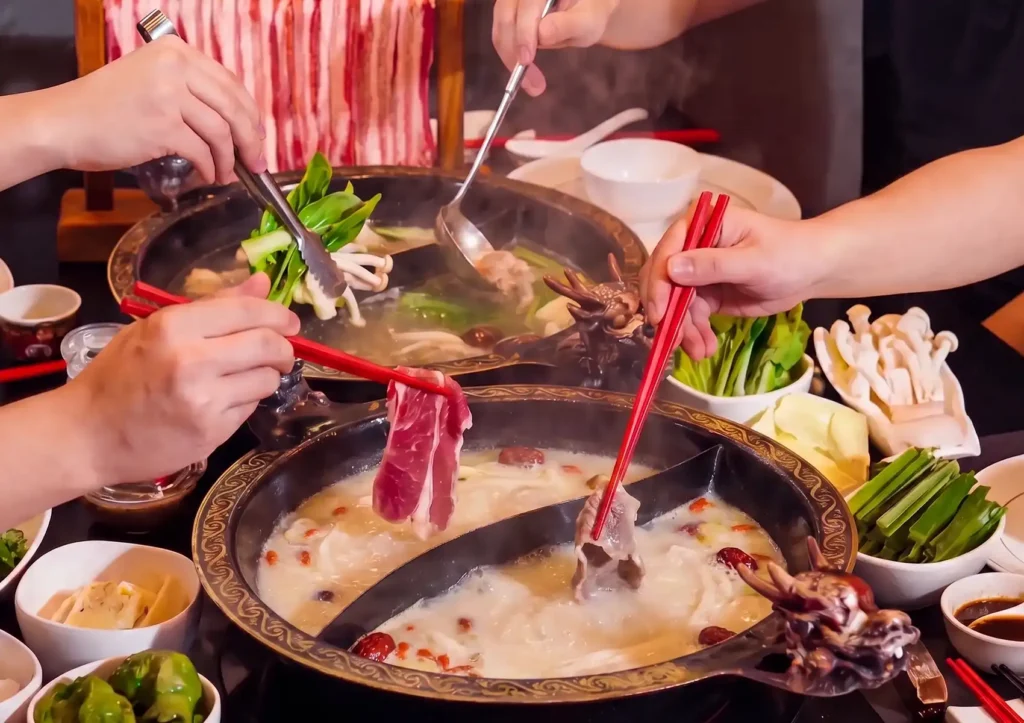 Diners using red chopsticks and tongs to dip thinly sliced meat and fresh vegetables into a steaming, ornate split-pot of rich collagen broth at a Cantonese restaurant in Bali