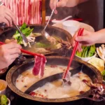 Diners using red chopsticks and tongs to dip thinly sliced meat and fresh vegetables into a steaming, ornate split-pot of rich collagen broth at a Cantonese restaurant in Bali