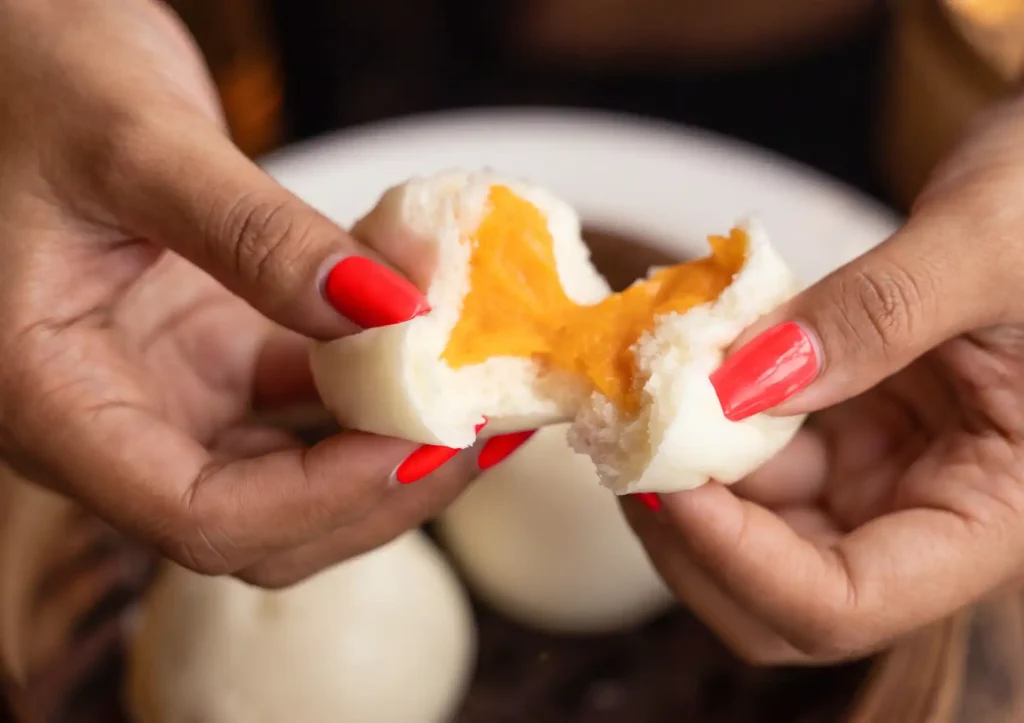 Two hands with bright red nail polish gently pulling apart a soft, fluffy steamed bun to reveal a warm, gooey orange custard filling, served fresh from a bamboo steamer at a Restaurant in ubud.
