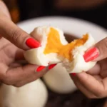 Two hands with bright red nail polish gently pulling apart a soft, fluffy steamed bun to reveal a warm, gooey orange custard filling, served fresh from a bamboo steamer at a Restaurant in ubud.