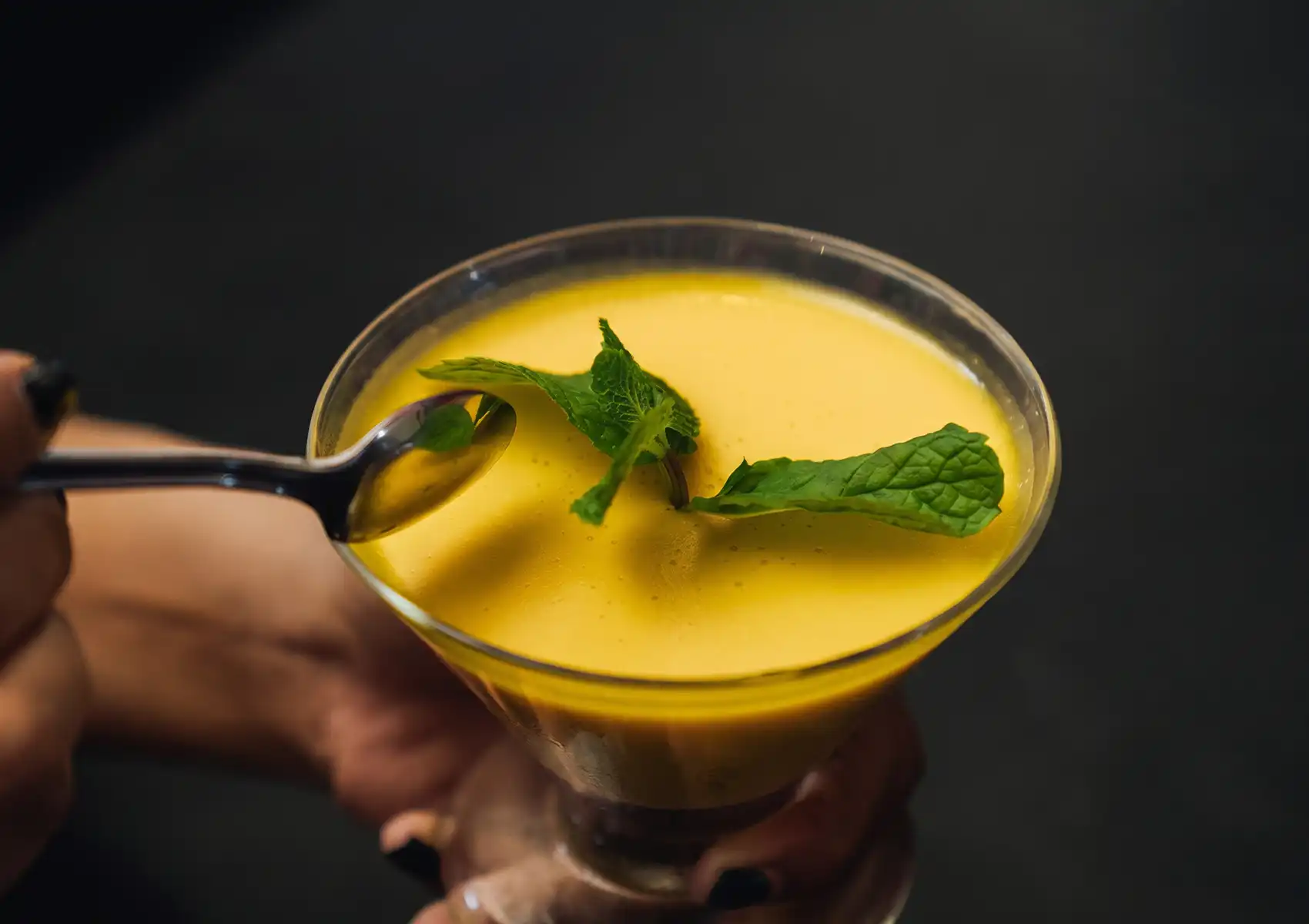 A close-up view of hands holding a glass dessert bowl filled with smooth, vibrant yellow mango pudding garnished with fresh mint leaves, preparing to take a spoonful at a Restaurant in ubud.