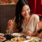 A smiling mother and daughter share a delightful meal of fresh dim sum, including steamed dumplings and siu mai, while seated in a cozy red booth at a Cantonese restaurant in Bali.
